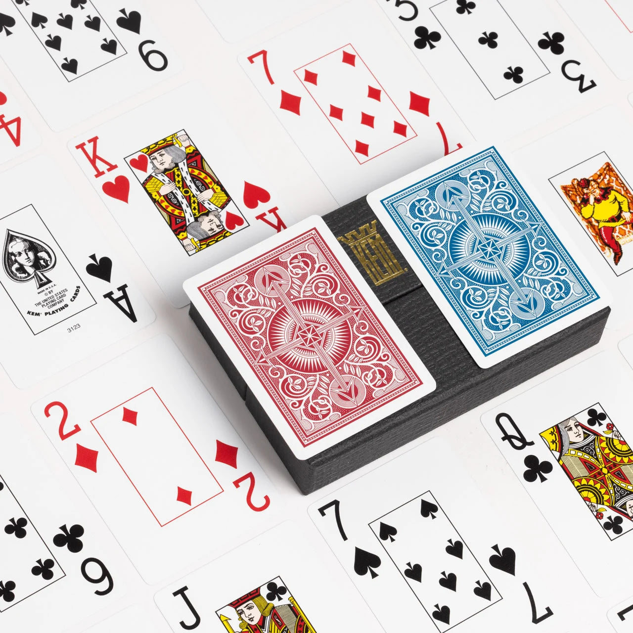 Two decks of KEM playing cards, one red and one blue, on a white background with scattered cards.