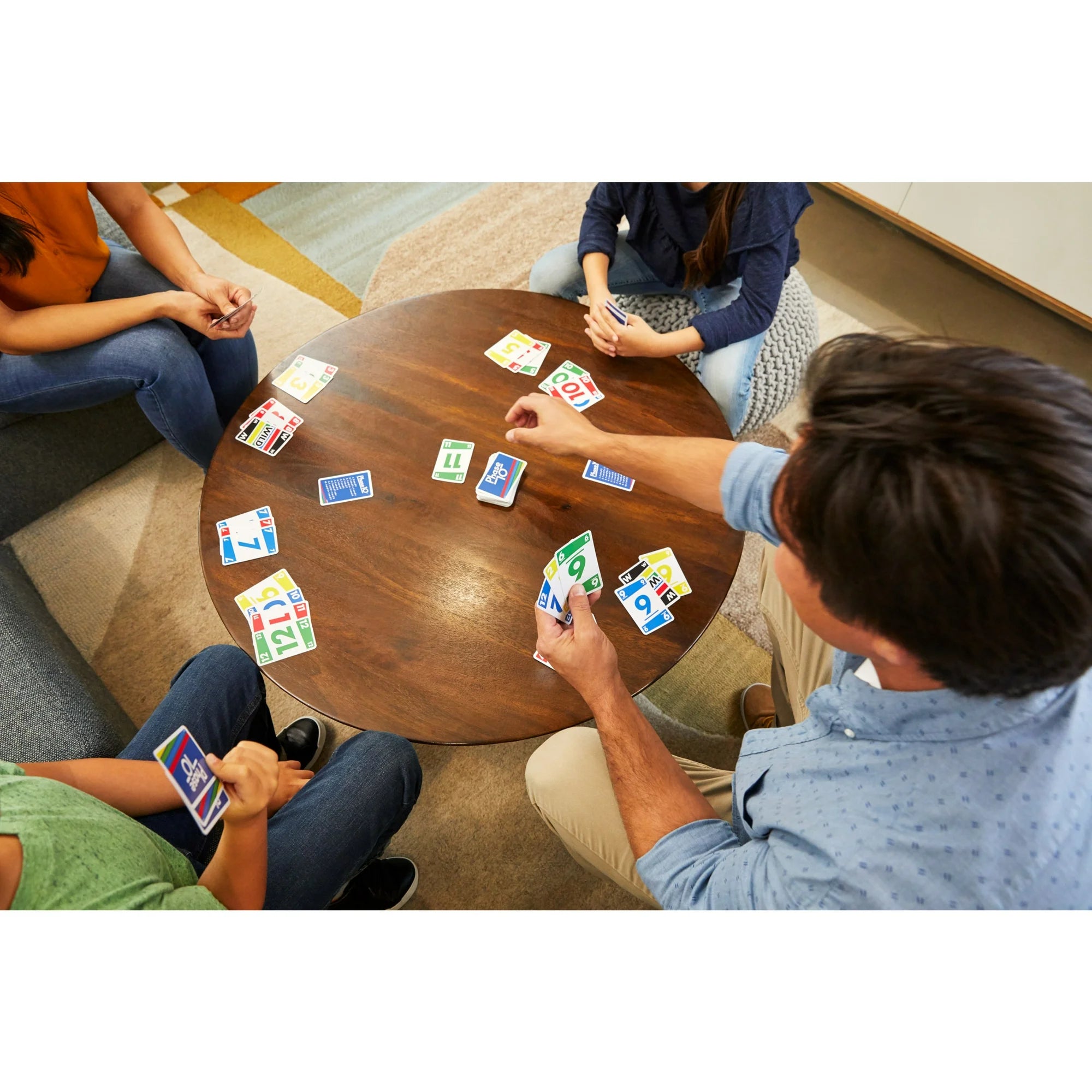 Four people sitting around a round wooden table playing Phase 10 card game in a modern indoor setting. The cards are spread across the table, and one person in the foreground is holding a hand of cards.