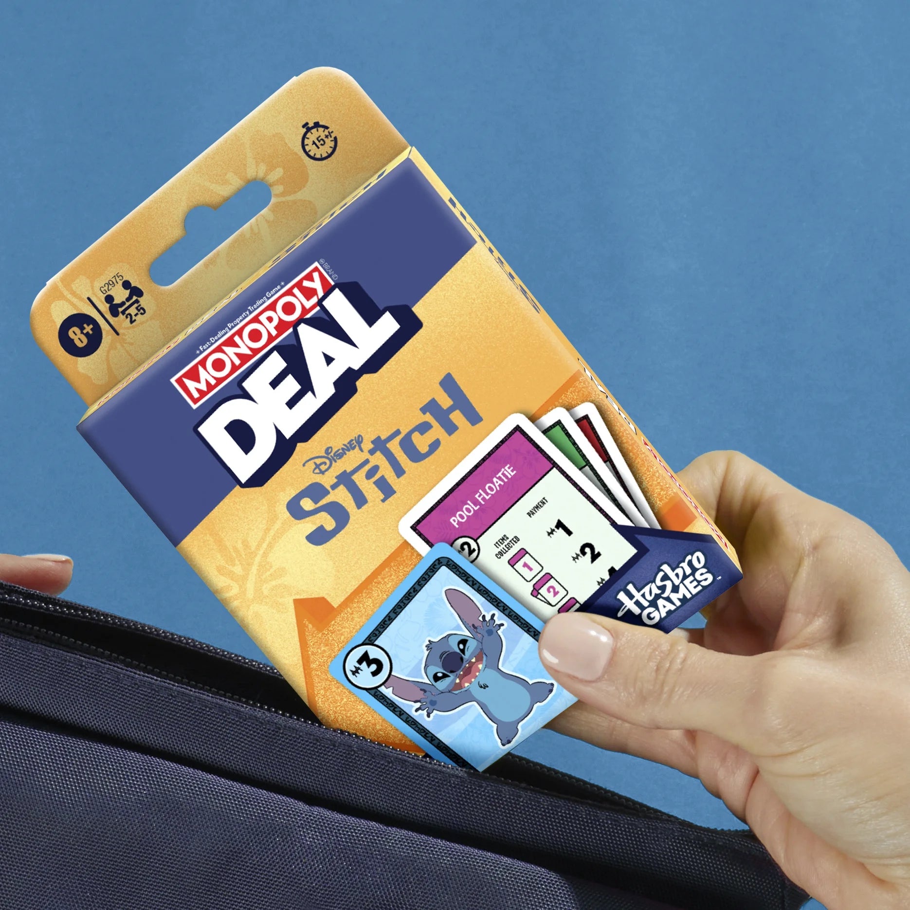 A hand holding a navy zippered pouch while sliding a Monopoly Deal Disney Stitch card game package and cards into the pocket, shown against a solid blue background.