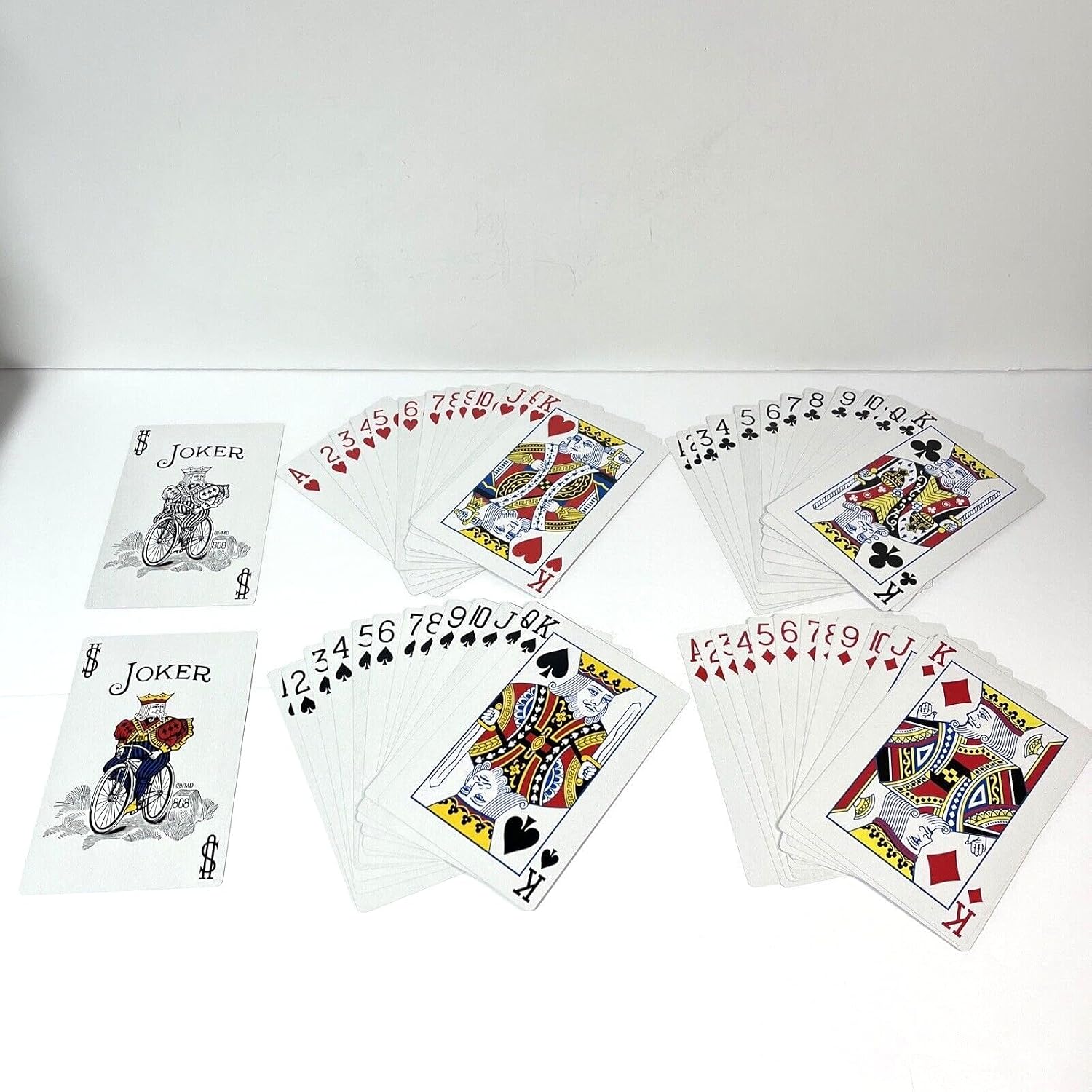 A set of Bicycle Big Box playing cards arranged on a white surface, including several face-up cards showing the Joker and Kings with fanned backs, and several face-down cards.