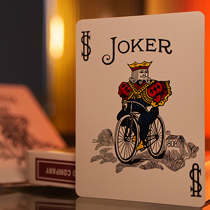 Joker card from a deck of pBicycle Metallic Ruby Playing Cards with a decorative design on a reflective surface.
