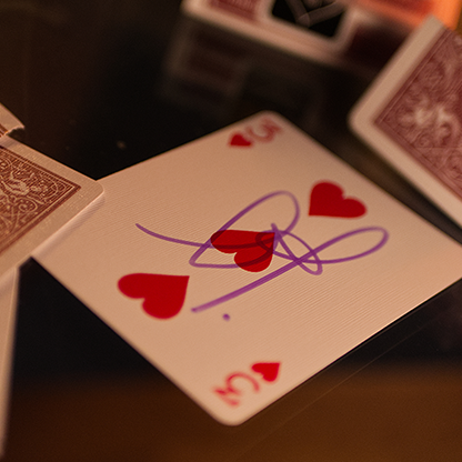 Bicycle Metallic Ruby Playing Cards with red hearts and a signature on a dark surface