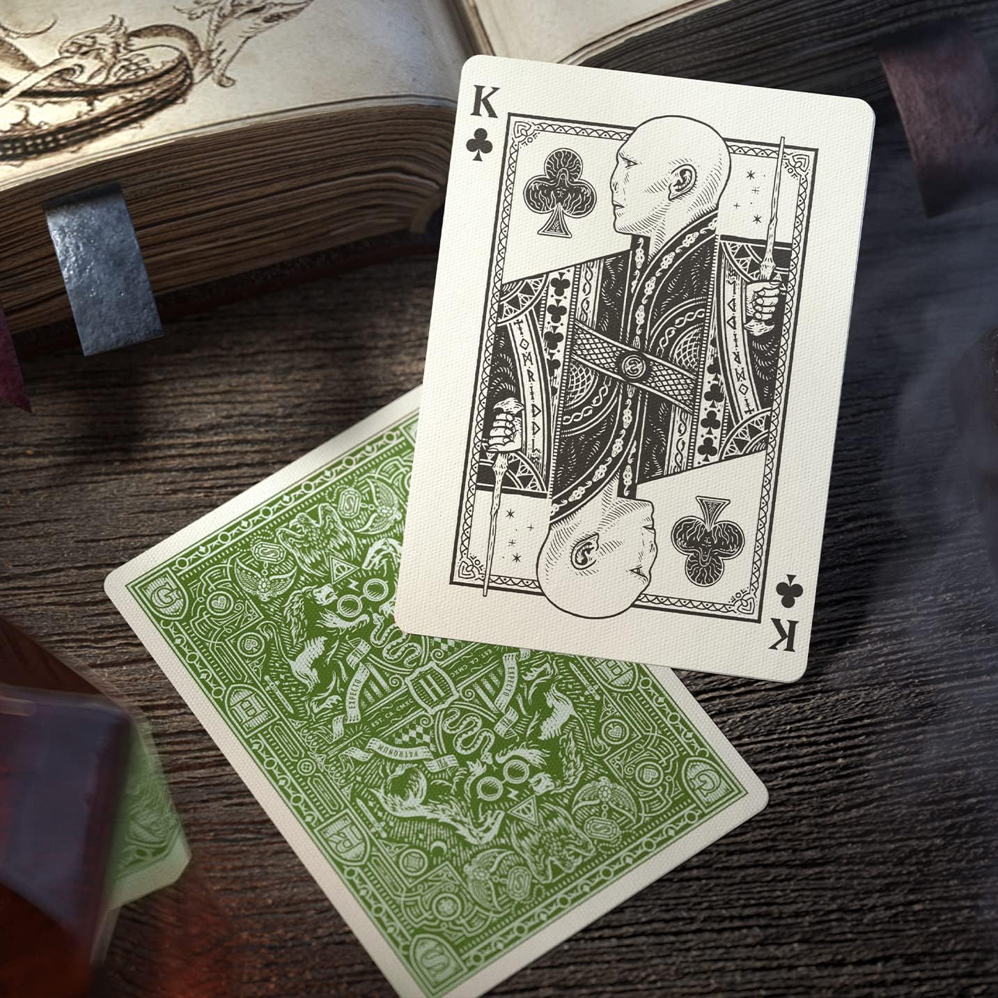 Two playing cards, King of Clubs and green back card, on a wooden surface with an open book.