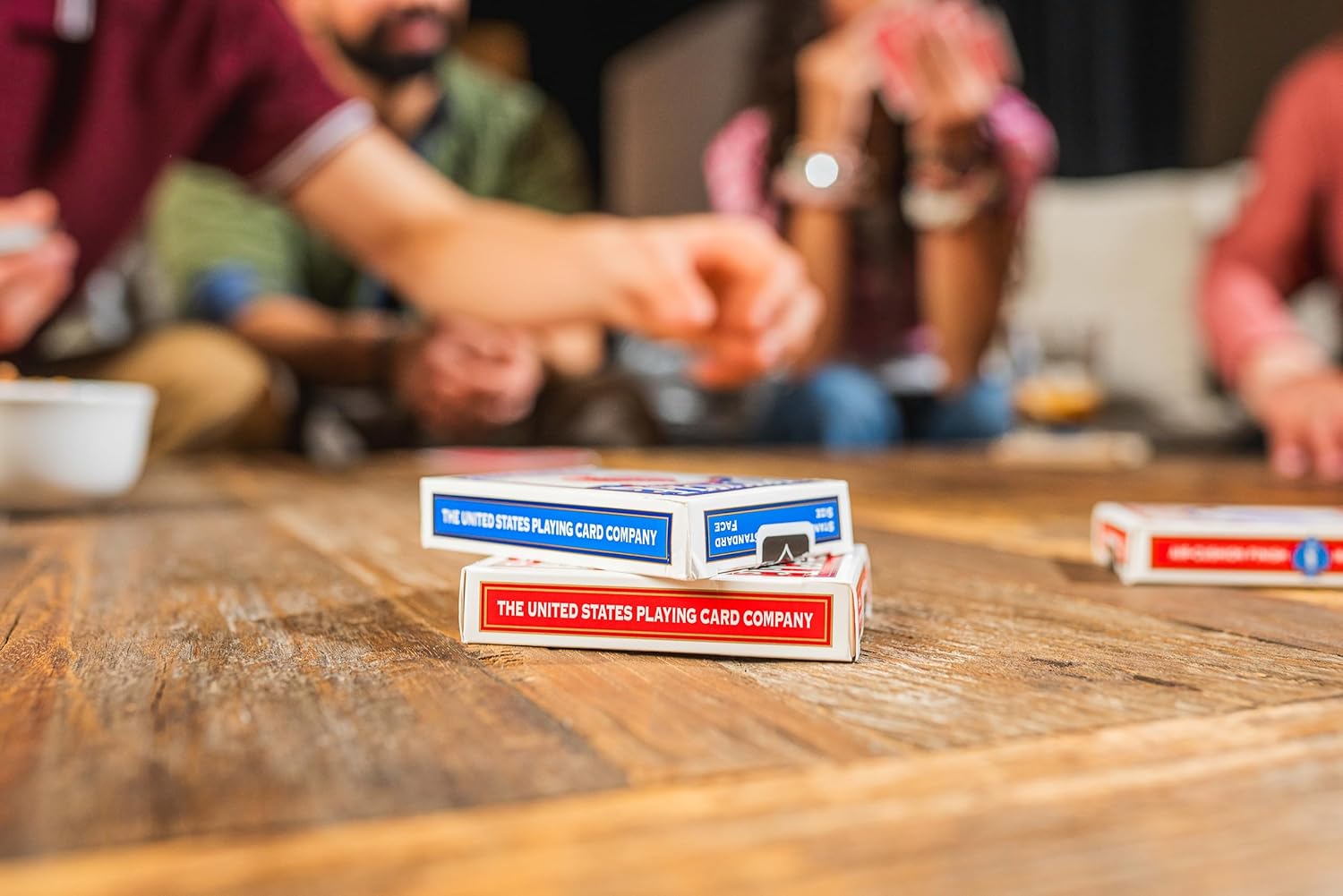 Two packs of United States Playing Card Company cards on a wooden table with people in the background.