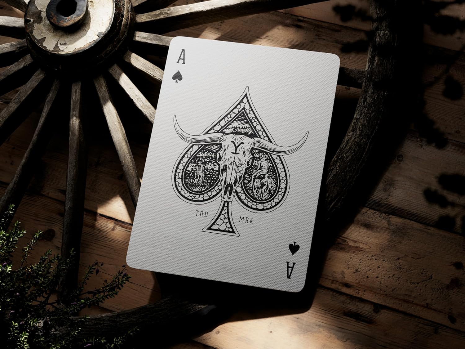 Playing card with ace of spades design featuring a bull skull, placed on a wooden surface with a rustic wheel in the background.