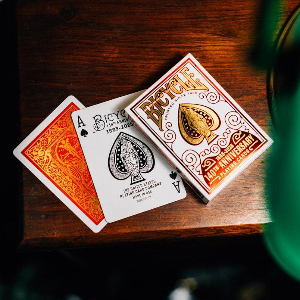 Bicycle 140th Anniversary playing cards on a wooden surface with a blurred green object in the foreground.