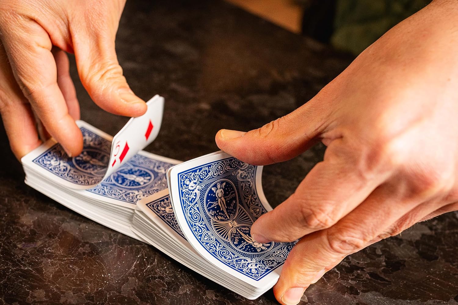 Close-up of hands shuffling a deck of blue and red playing cards on a dark surface.