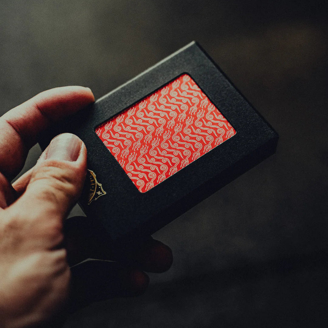Hand holding a black box with a NO BORDERS: Series 2 Playing Cards red crown patterned card inside against a dark background