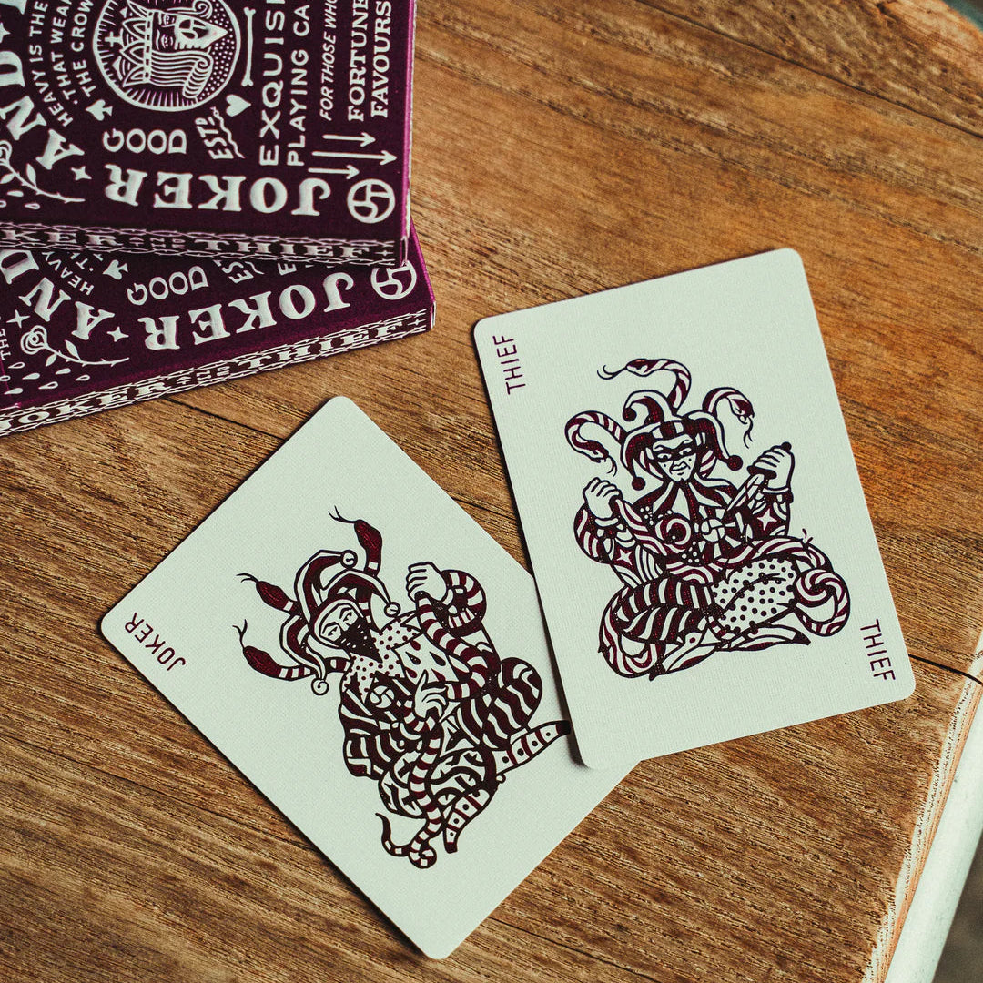 Two Crown: Royal Purple Edition playing cards with intricate designs on a wooden surface, with a deck of cards in the background.