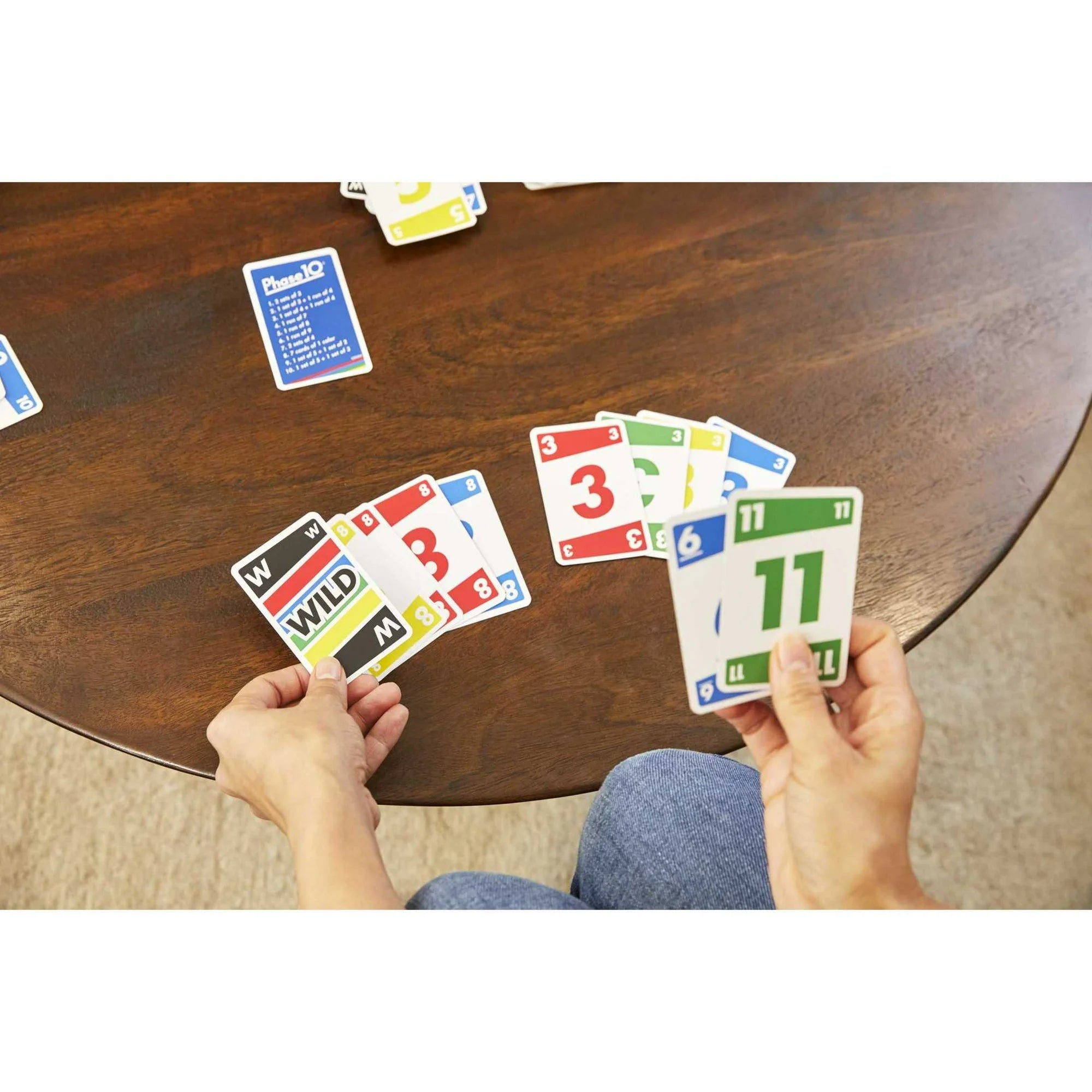 A top-down view of a person playing Phase 10 card game at a round wooden table, holding two fanned-out cards in both hands. The cards have colorful borders and numbers, and other cards are scattered on the table.