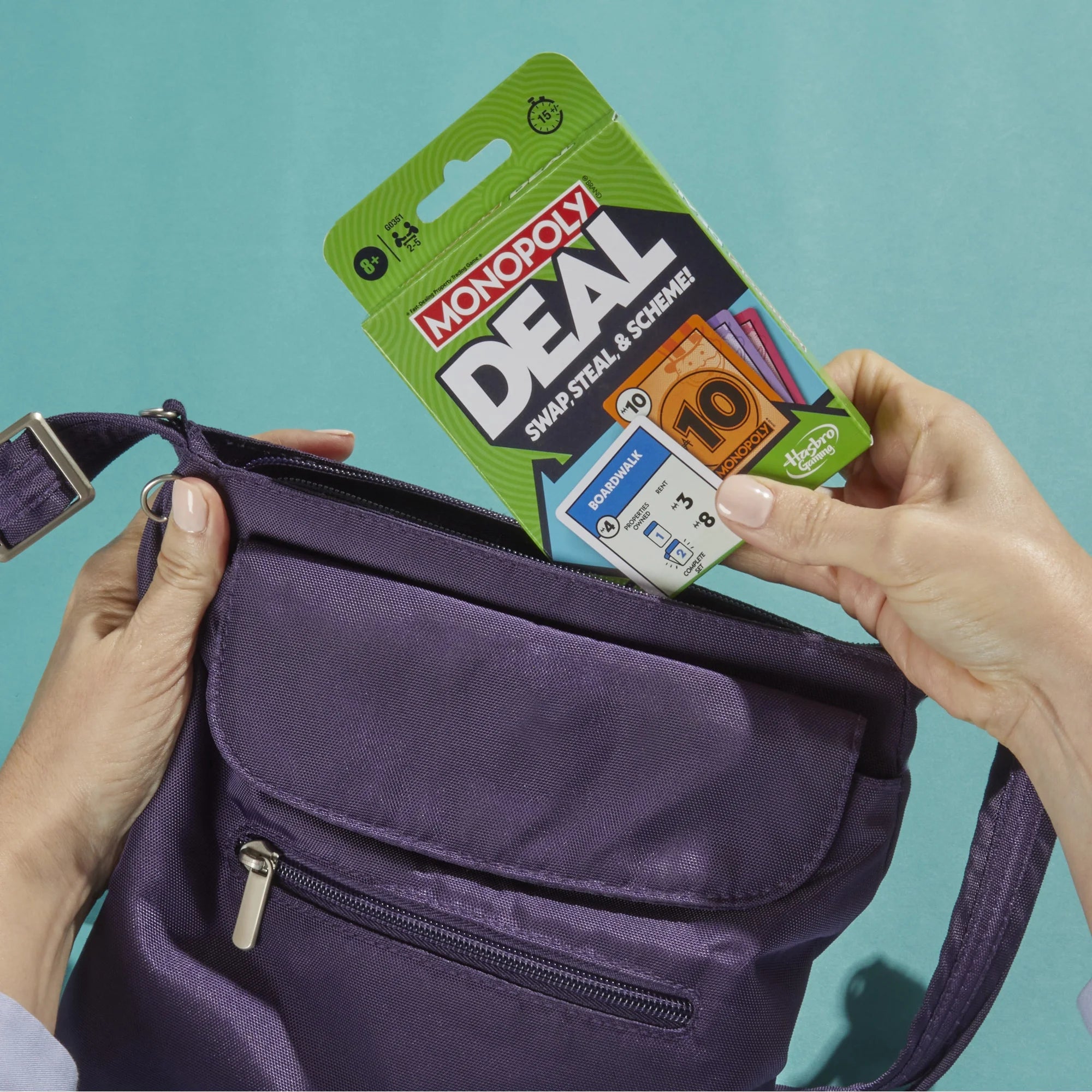 A purple nylon crossbody bag is shown with a Monopoly Deal card pack being inserted into its front zip pocket. Two hands are visible holding the bag and the card pack against a solid teal background.