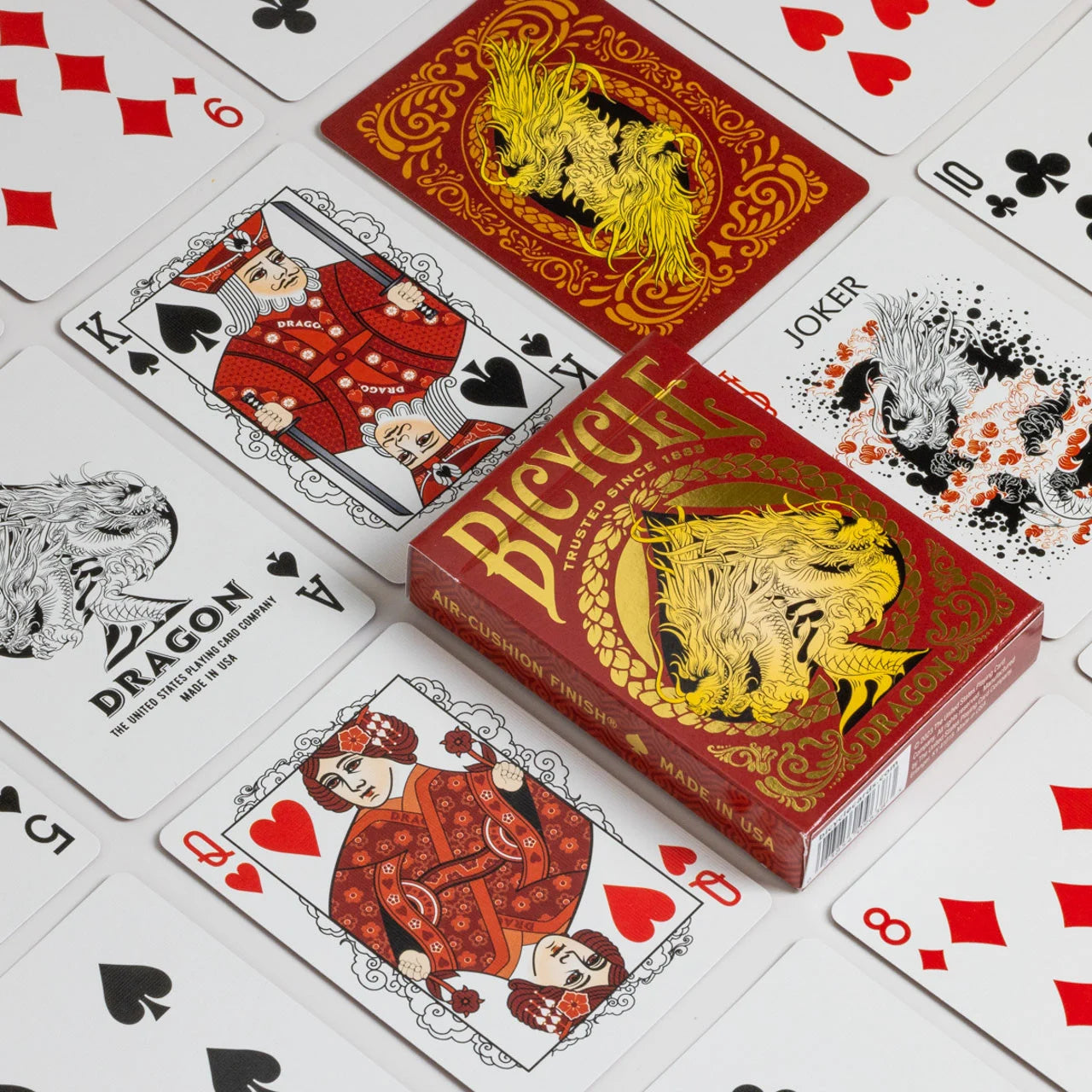 Bicycle Red Dragon deck spread out with custom dragon backs, red‑garbed courts, and Jokers surrounding the foiled tuck box.