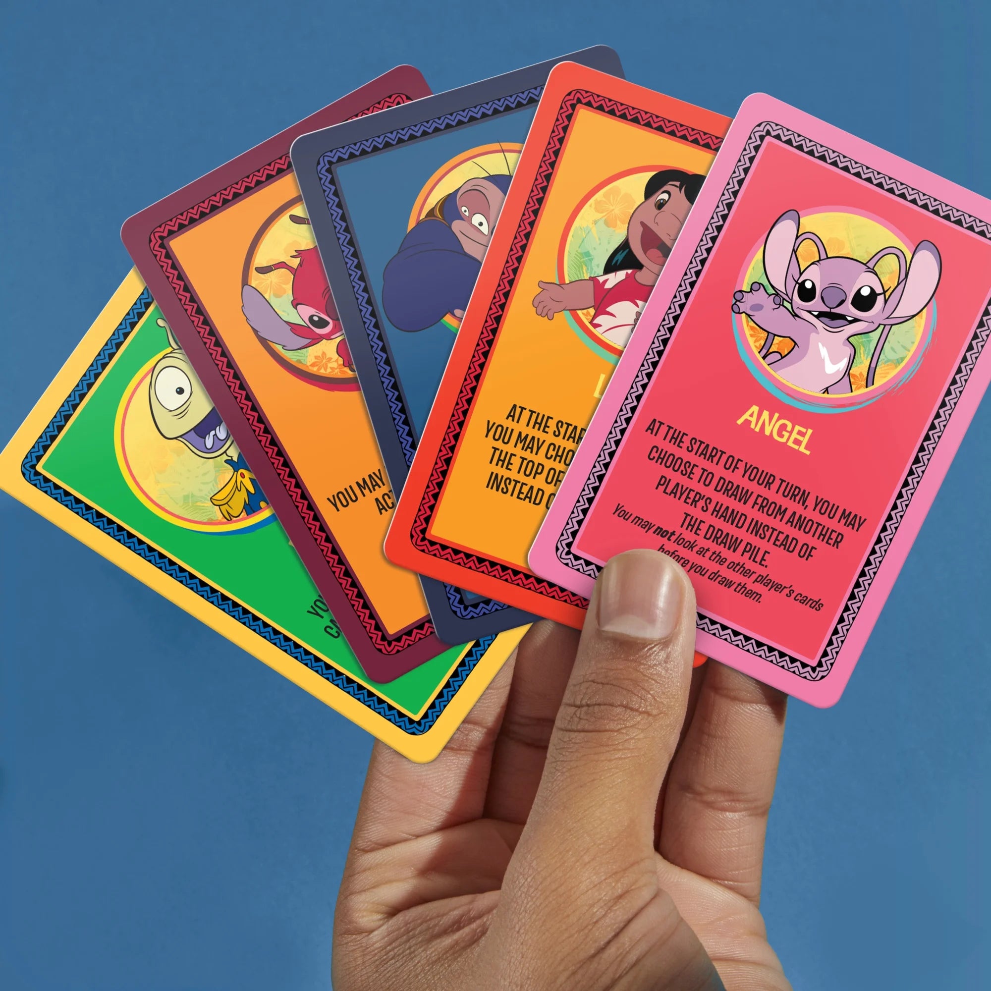 A hand holds five colorful Monopoly Deal Disney Stitch character cards against a solid blue background. The cards feature bright borders and character illustrations, including Angel from Lilo & Stitch.