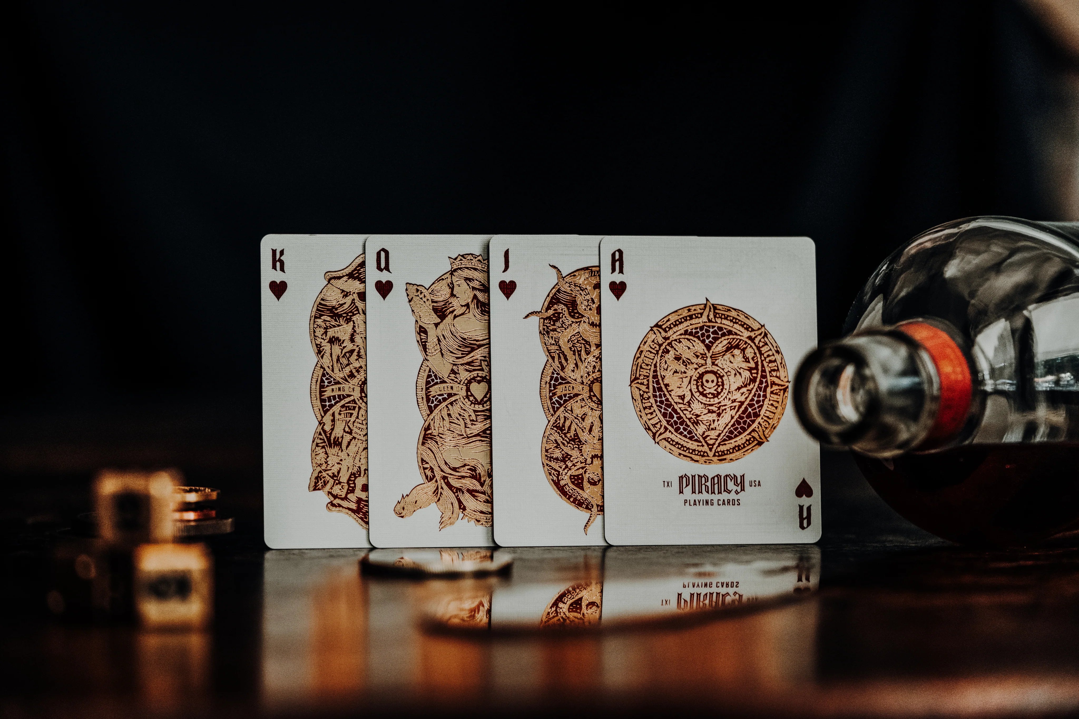 Four Piracy playing cards with intricate designs on a reflective surface next to a bottle.
