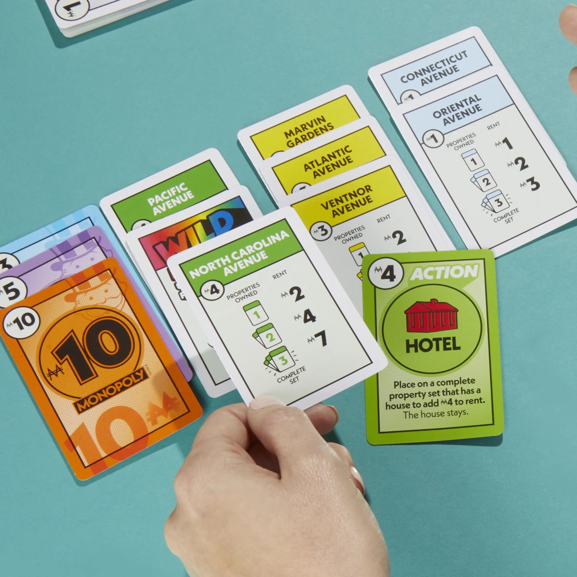 Close-up of Monopoly Deal game cards on a teal surface with a hand holding a card.
