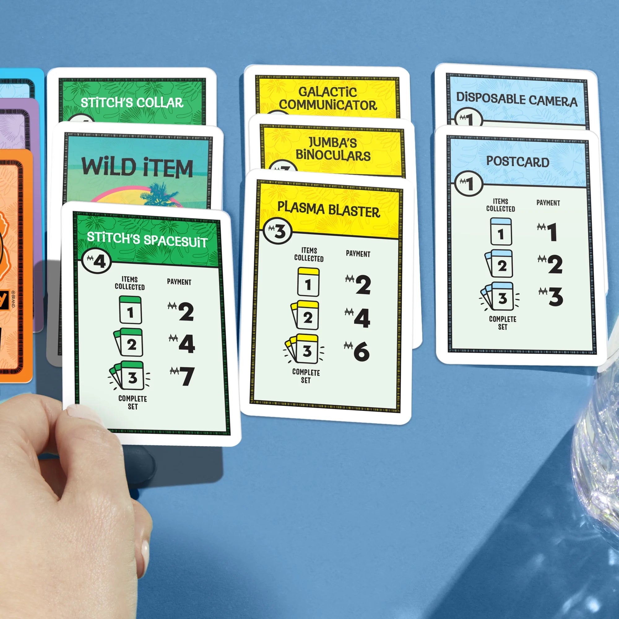 Collection of Monopoly Deal Disney Stitchcards on a blue surface with a hand holding one card.