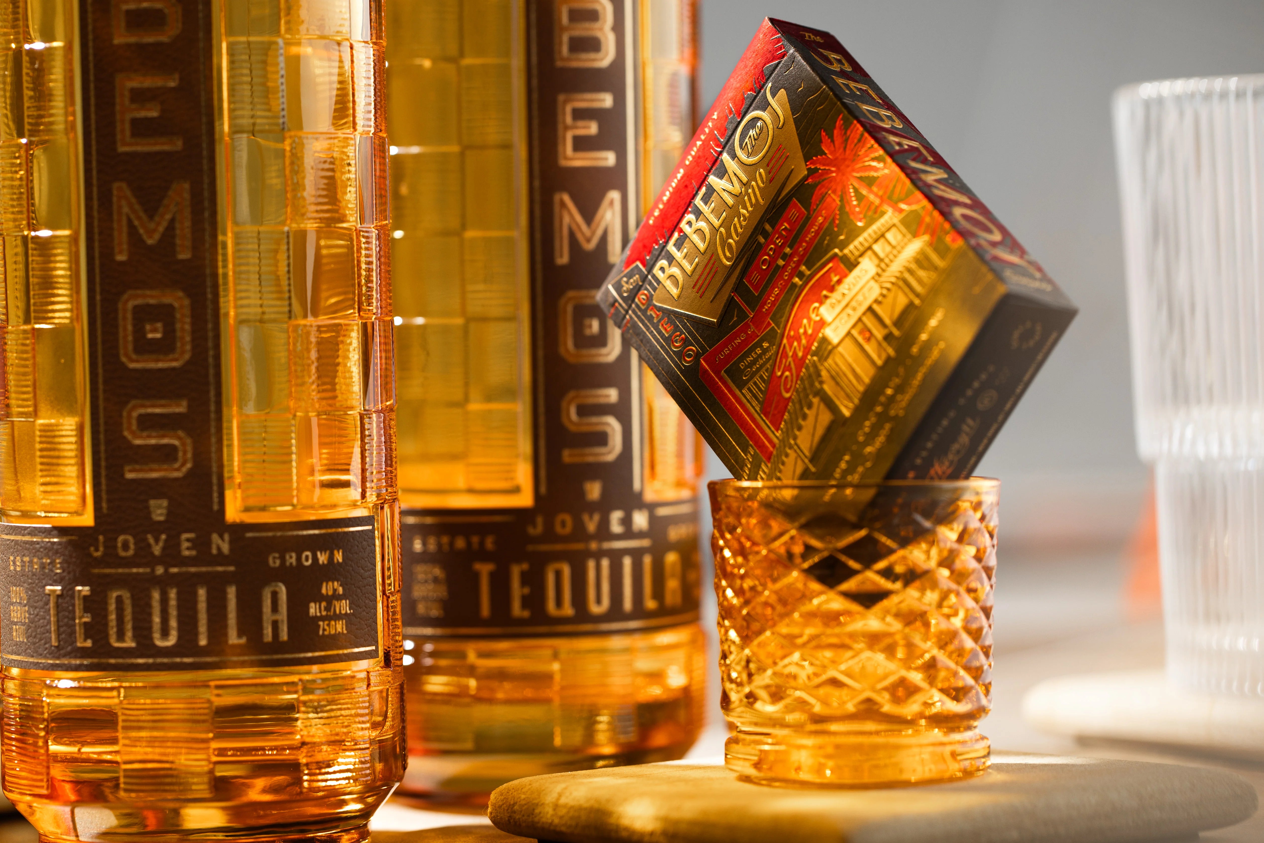 Two bottles of Remos Joven Tequila with a decorative glass and box of Bebemos Casino playing cards on a light background.