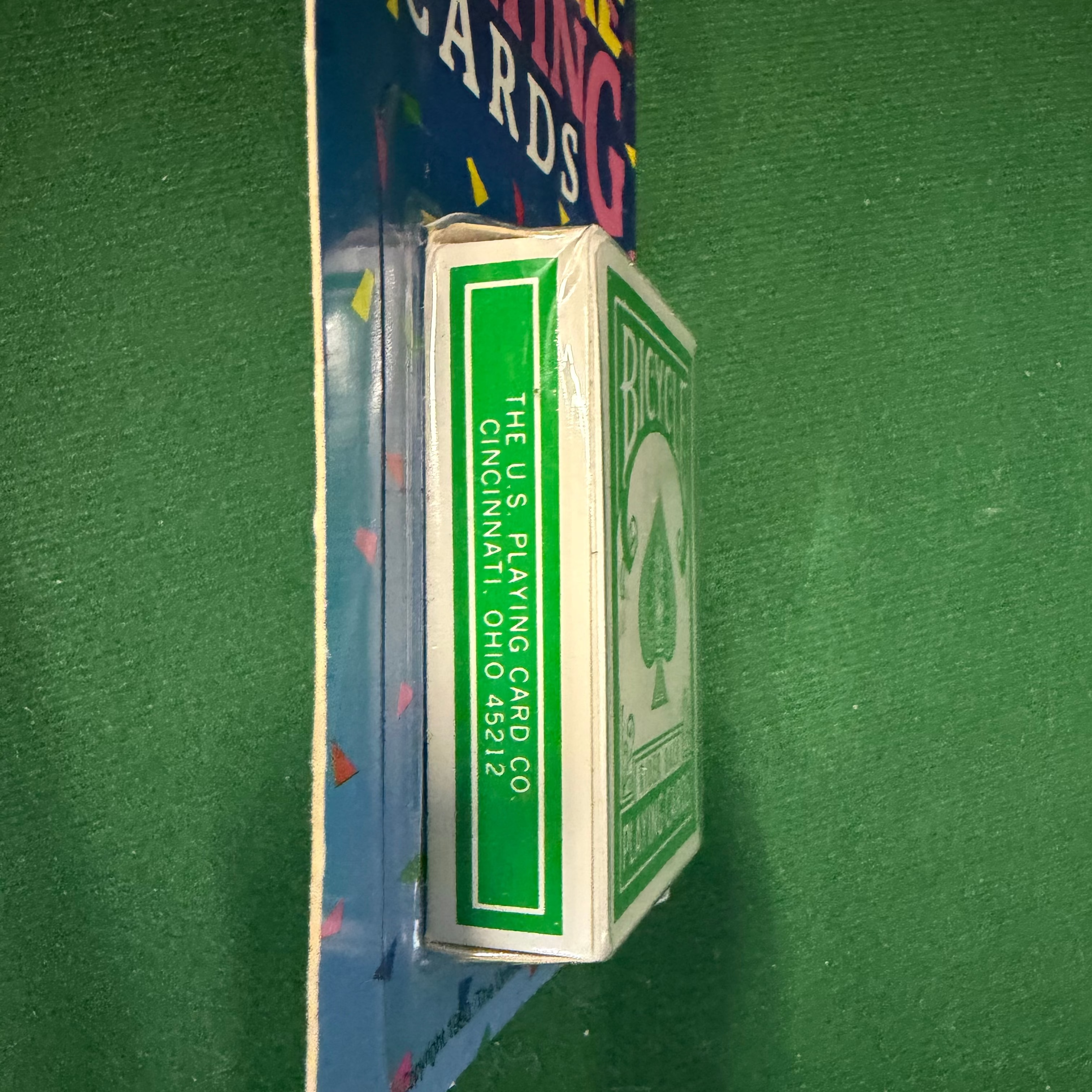 A deck of playing cards in its original blue and white cardboard packaging, standing vertically on a green felt surface.