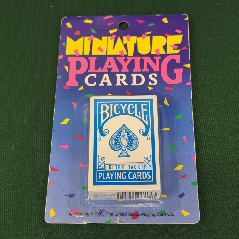 A package of miniature blue Bicycle Rider Back playing cards in clear plastic blister packaging on a purple card backing with colorful confetti graphics from 1990, placed on a green textured surface.