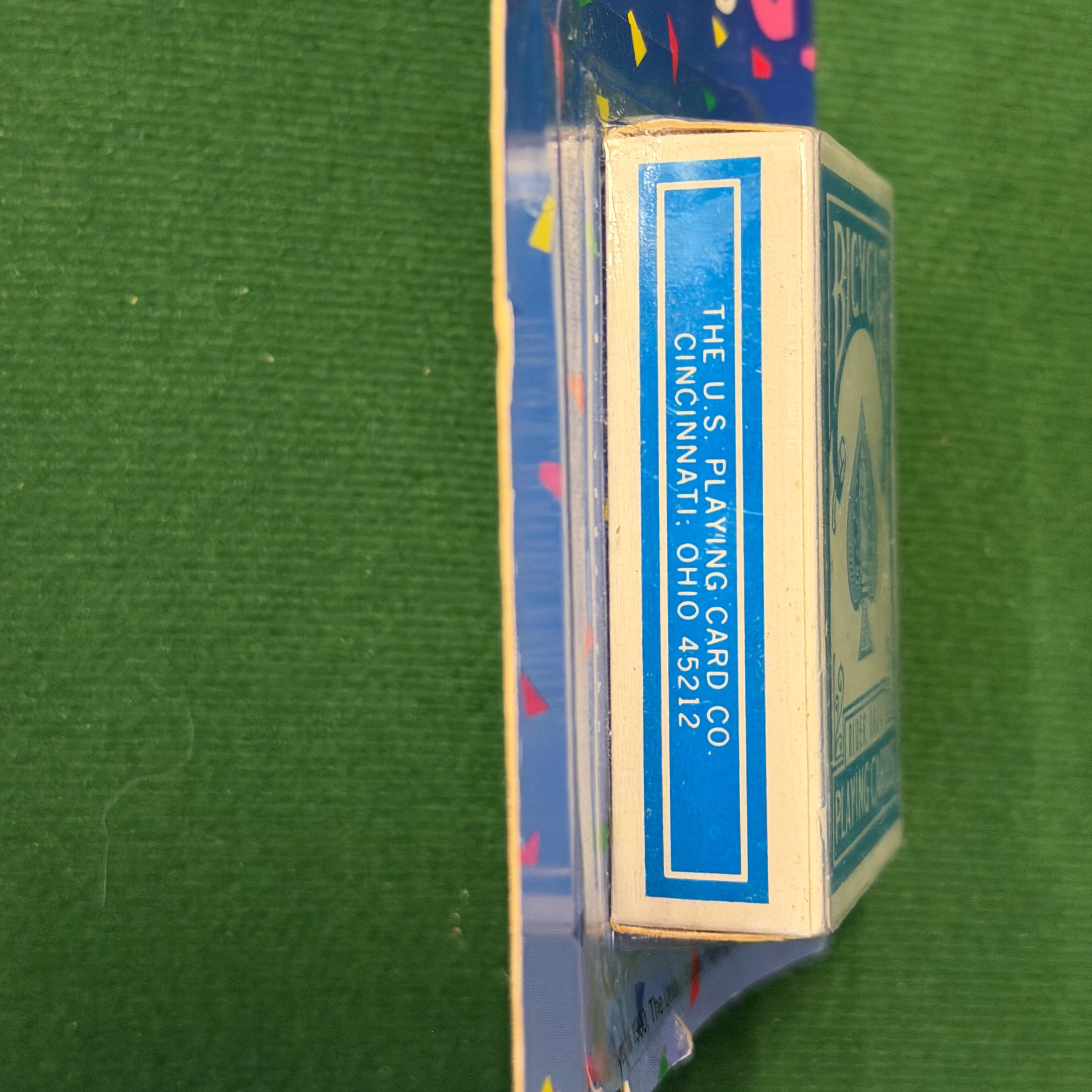 A vintage deck of playing cards in its original blue cardboard packaging, standing vertically on a green felt-like surface. The side of the box shows 'THE U.