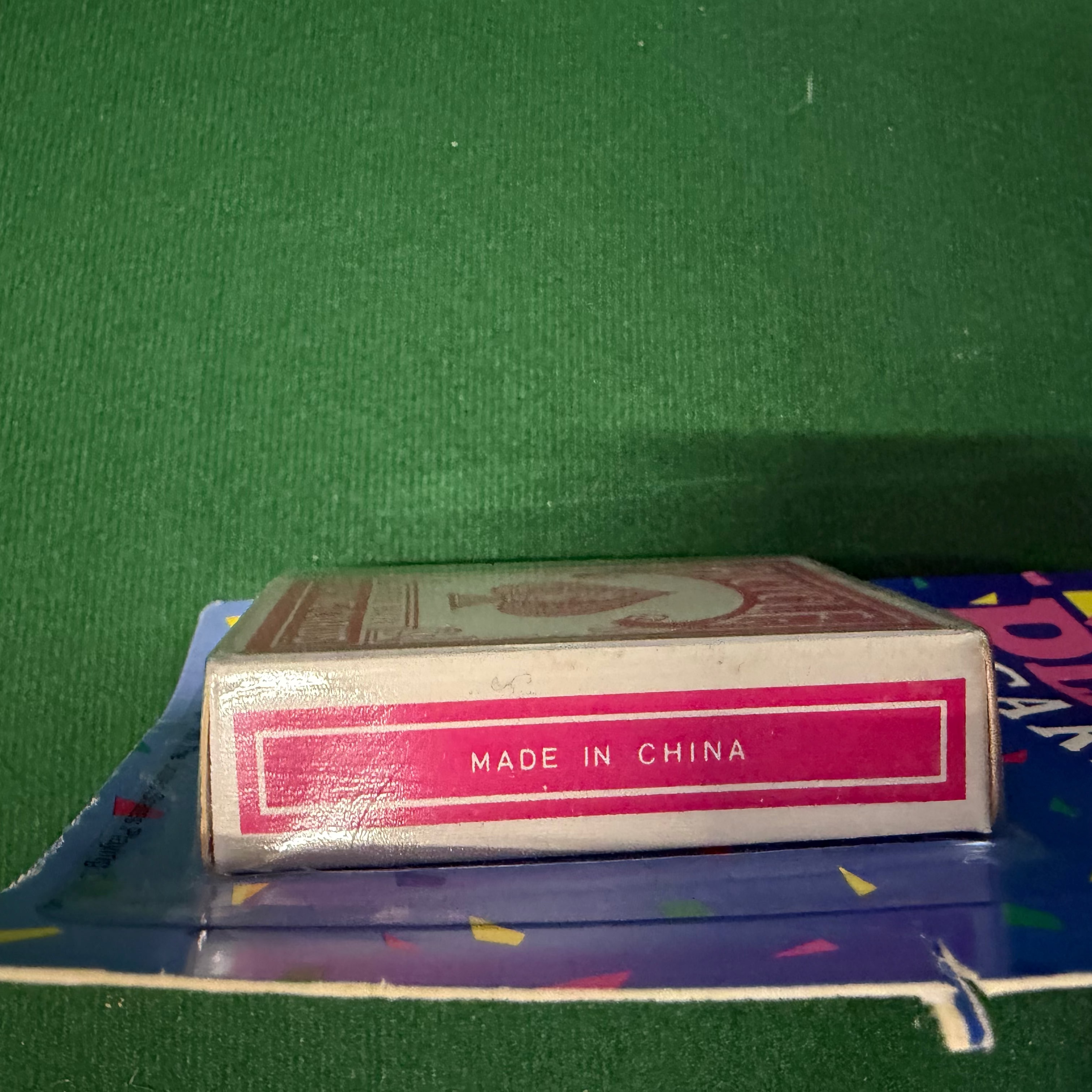 A vintage deck of playing cards in a clear plastic sleeve, showing the spine with 'MADE IN CHINA' printed in white on a magenta band. The deck rests on a blue card backing against a green felt-like surface.