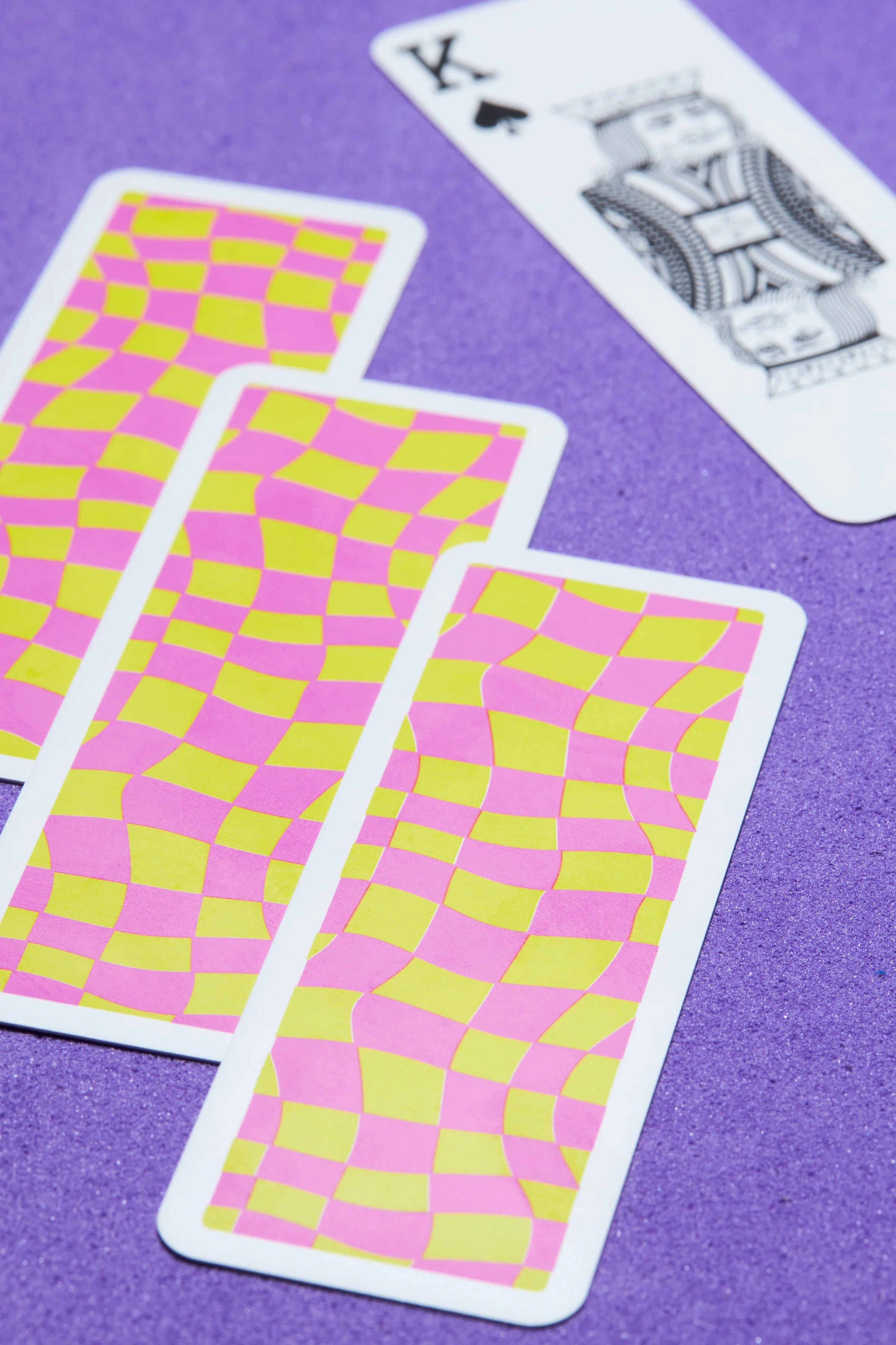 Three Copag Go Deck – Chess Design playing cards with pink and yellow pattern on a purple surface, one card is face up showing a king of spades.
