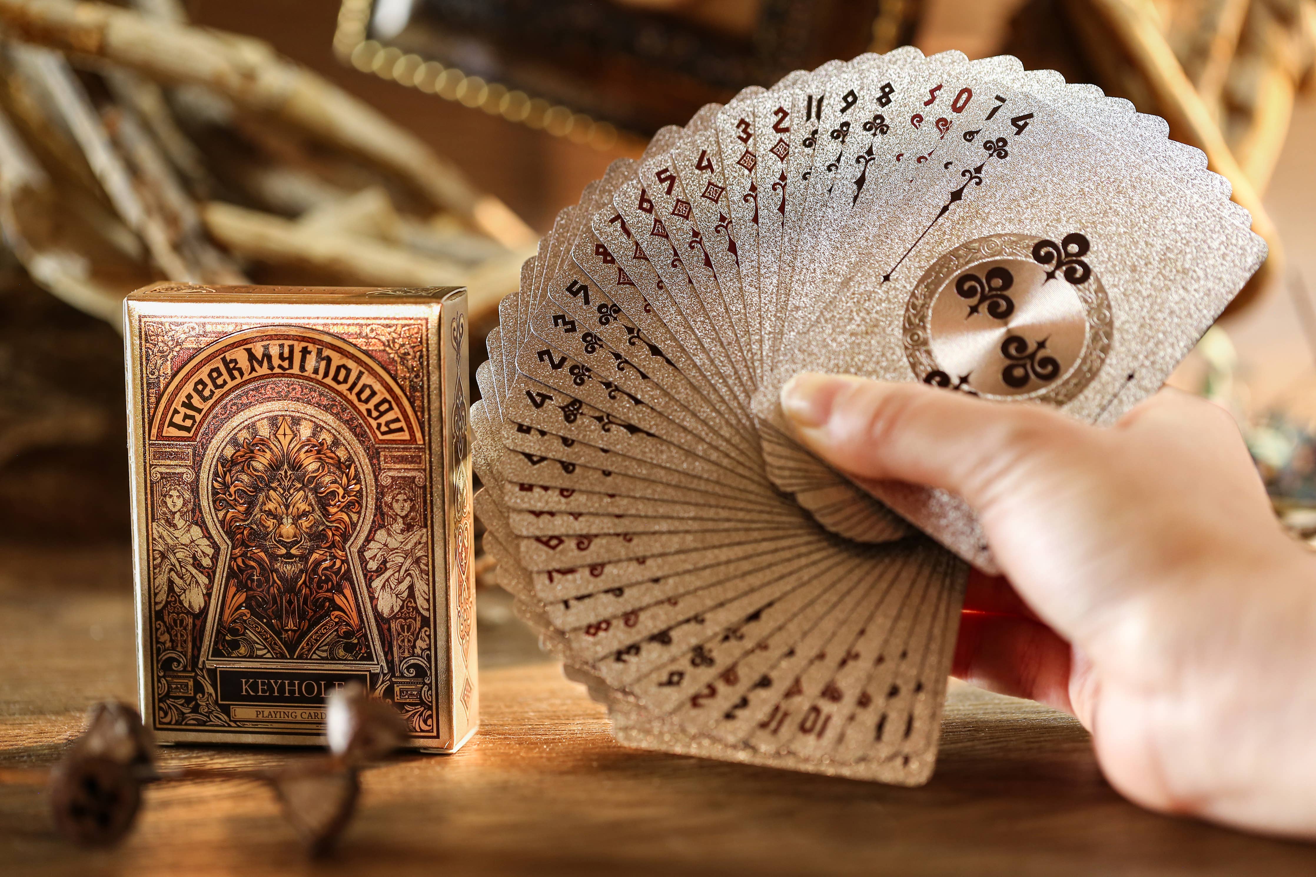 Hand holding a deck of cards next to a 'Greek Mythology' playing card box on a wooden surface.