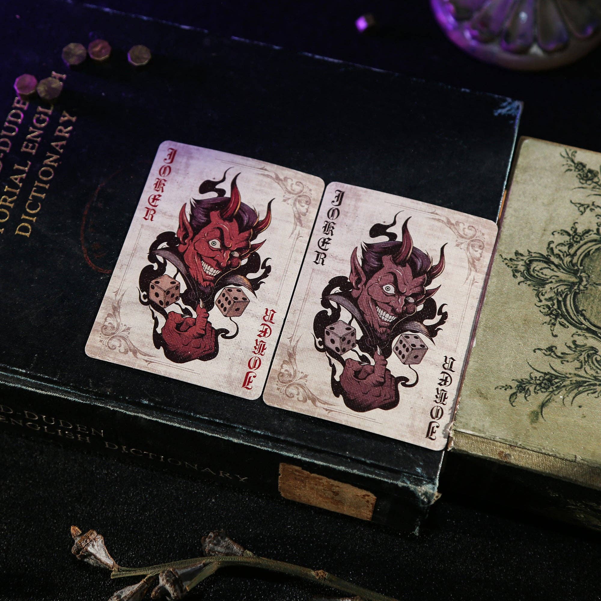 Two playing cards with devil designs on a dark surface with an open book.