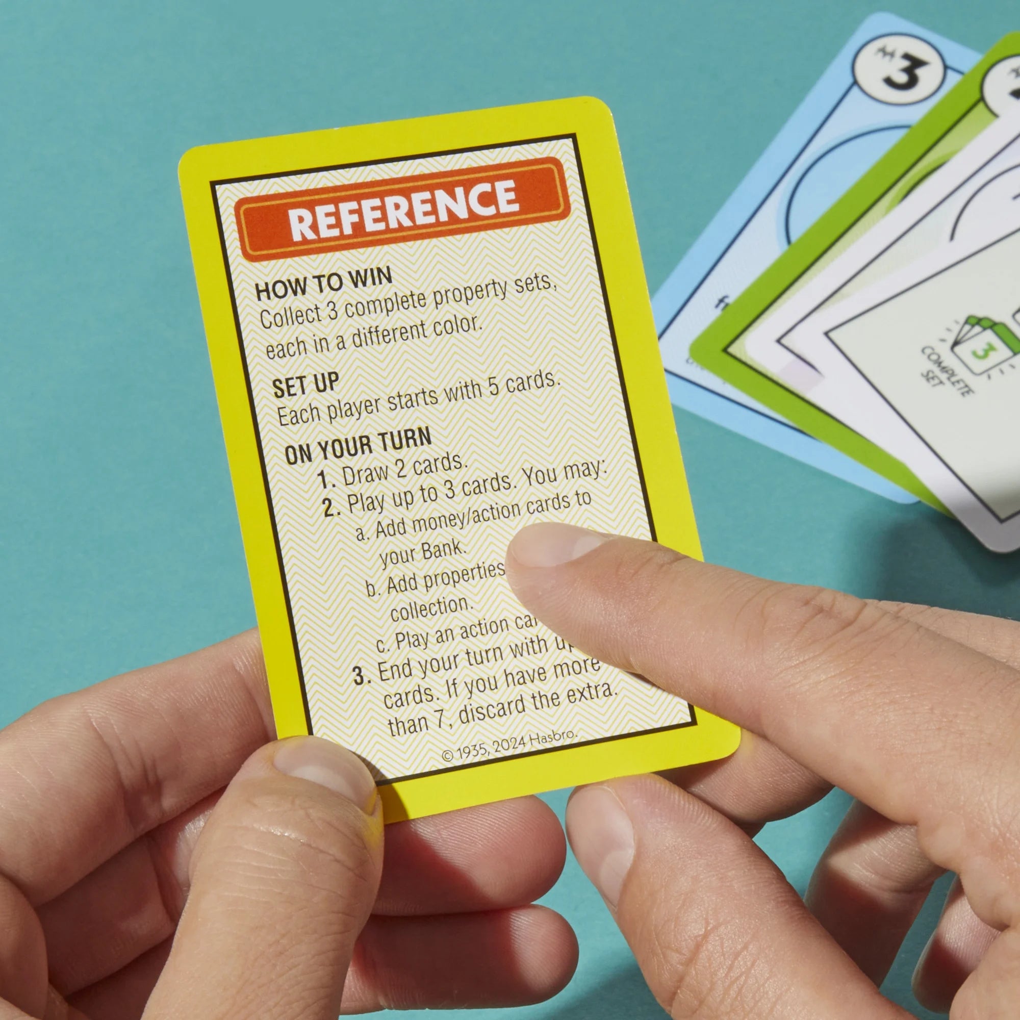 Hand holding a Monopoly Deal card with game rules against a teal background