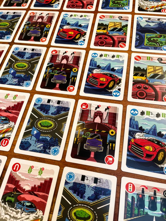 A close-up view of a Driving Me Crazy: Route 66 100th Anniversary Card Game
 with multiple illustrated cards arranged in a grid on a brown surface. The cards feature stylized cars, roads, and city scenes in bright colors, with red, blue, green, yellow, and black artwork.