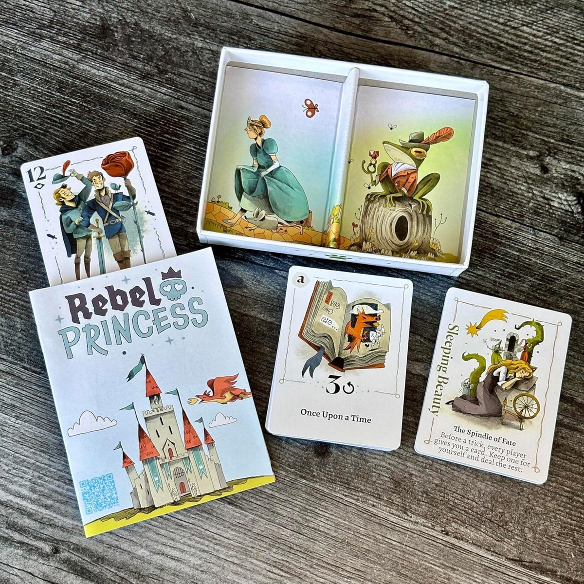 Rebel Princess standard edition box, illustrated playing cards, and round cards on a wooden table.