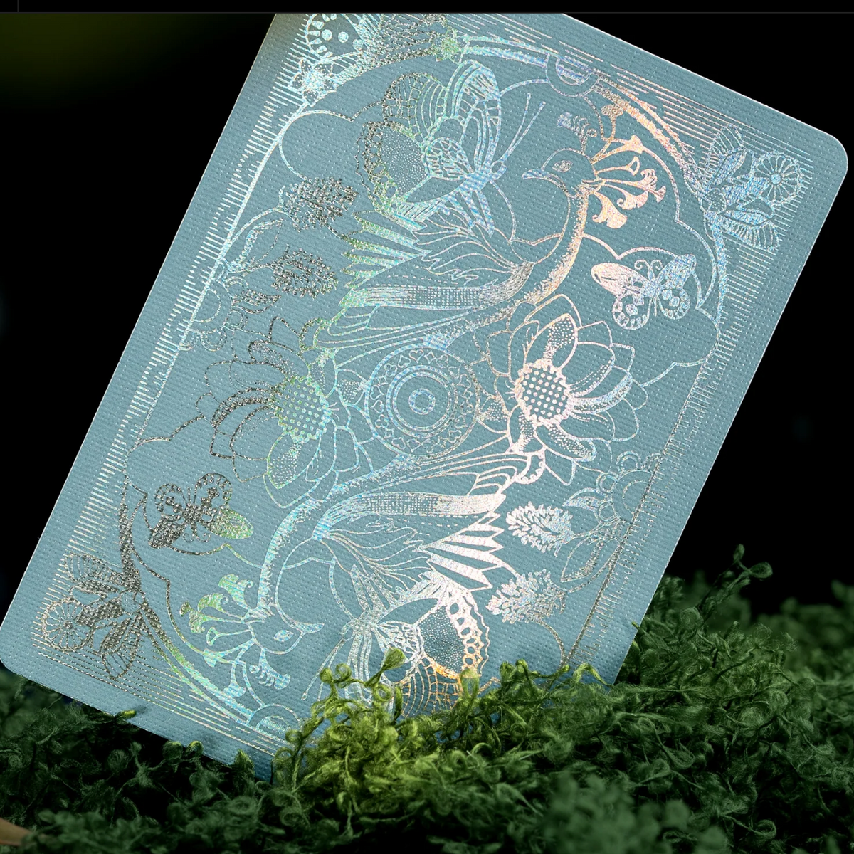 Light blue Memento Moooi card back with holographic foil birds, flowers, and butterflies, displayed among green moss.