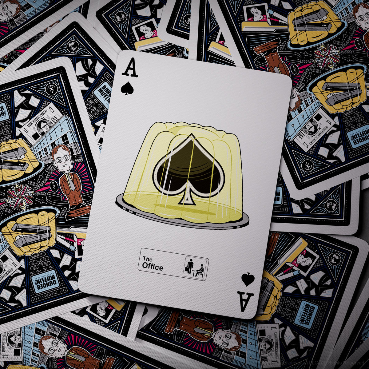 The Office Playing Cards Ace of Spades with a stapler-in-gelatin illustration over scattered card backs